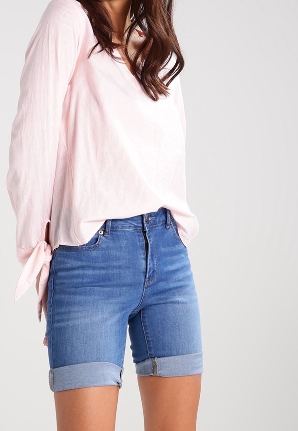 Chemise rose clair à manches longues avec liens sur les côtés, associée à un short en jean bleu avec revers roulés et taille mi-haute.