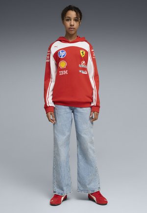 Jong persoon staand, gekleed in een rode en witte Ferrari-trui met sponsormerken, lichtblauwe jeans en rode sneakers tegen een neutrale achtergrond.