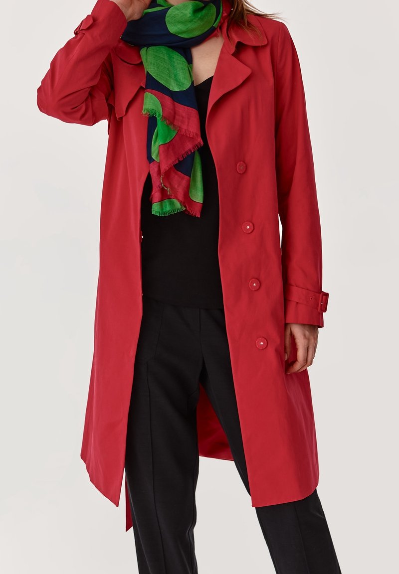 Mujer vestida con un abrigo de trinchera rojo con botones, blusa y pantalones negros, y una bufanda colorida con patrones circulares en verde, azul marino y rojo.