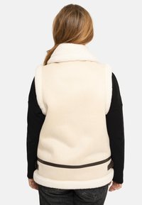 Personne aux cheveux longs portant un gilet sans manches en peau lainée couleur crème par-dessus une chemise noire à manches longues, vue de dos sur un fond blanc.