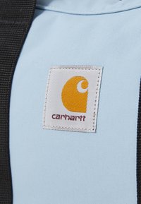 Tissu bleu clair avec une étiquette carrée blanche présentant un logo jaune et un texte rouge "carhartt". Des sangles noires sont visibles sur le côté.