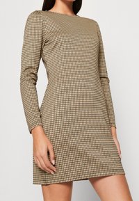 Robe à manches longues en tissu beige avec un motif en grille noir, silhouette ajustée, encolure ronde et texture douce. Ourlet au-dessus du genou.
