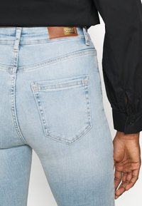 Pantalones vaqueros de denim azul claro con forma ajustada, que cuentan con bolsillos traseros y un parche de cuero marrón en la cinturilla. Textura suave.