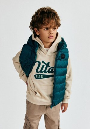 Teal pufīgi vestes virsū krēmkrāsas kapuces džempera ar zaļiem uzrakstiem, kopā ar bežām kargo biksēm, uz neitrāla fona.