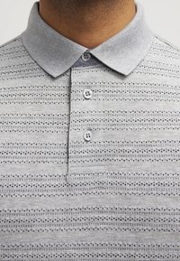 Polo gris avec un tissu texturé comportant des rayures horizontales et de petits motifs. Patte de boutonnage à trois boutons avec un col contrasté.
