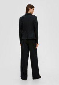 Vrouw met bruin haar draagt een zwarte blazer en wijde broek, staand met de rug naar de camera tegen een effen lichtgrijze achtergrond.