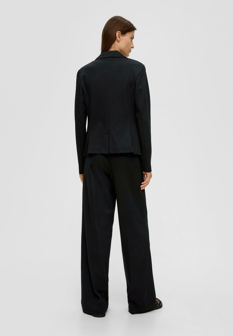 Femme aux cheveux bruns portant un blazer noir et un pantalon large, se tenant de dos devant un fond gris clair uni.