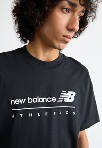 Černé bavlněné tričko s bílým logem a textem "new balance ATHLETICS" v tučném, moderním písmu, s klasickým kulatým výstřihem a krátkými rukávy.
