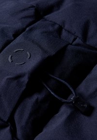 Tela de chaqueta acolchada azul marino con una textura suave, que presenta un logo circular y un cordón con un tope de plástico para realizar ajustes.