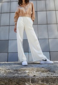 Hellbeige Sweatshirt mit halbem Reißverschluss und weißen Jogginghosen. Weiße Sneakers mit schwarzen Akzenten. Glattes Stoffgefühl, lockere Passform.