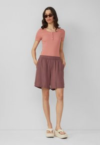 Jasnoróżowa, dopasowana bluzka z krótkim rękawem i guzikami, połączona z luźnymi, sięgającymi kolan fioletowo-różowymi szortami oraz beżowymi sandałami na platformie.