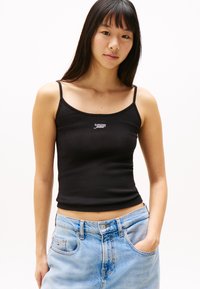 Black cotton tank top with thin straps and a fitted design. Features a small white "Tommy Jeans" logo on the chest. Paired with light blue jeans.