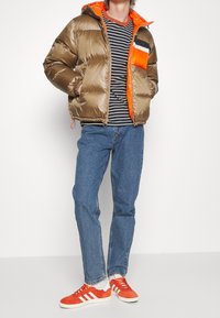 Personne portant une doudoune marron brillante avec une doublure orange, un t-shirt rayé noir et blanc, un jean bleu, et des baskets orange vif avec des rayures blanches.