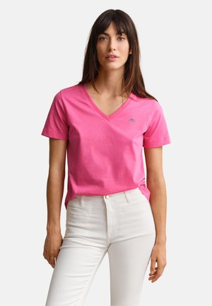 Woman wearing a bright pink V-neck short-sleeve shirt and high-waisted white pants, standing against a plain light background.
