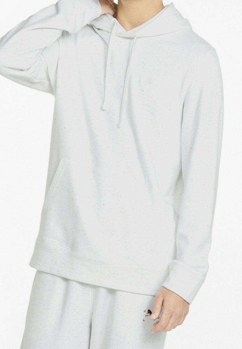 Persona con una sudadera con capucha gris claro moteada y pantalones de chándal a juego, con un pequeño logo en el muslo izquierdo, mano levantada cerca del cuello.