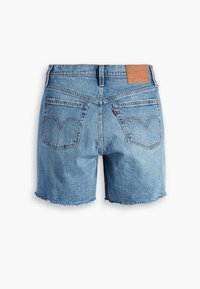 Short en jean bleu clair avec ourlets effilochés, deux poches arrière avec détails de couture, passants pour ceinture et écusson en cuir beige sur la ceinture.