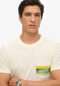 Camiseta de manga corta de color crema hecha de una tela texturizada. Presenta un bolsillo en el pecho con rayas verdes y amarillas y el estampado "Superdry".