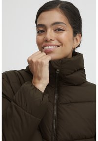Braune gepolsterte Jacke mit hohem Kragen und Reißverschluss, mit genähten Einsätzen und elastischen Bündchen. Das Modell passt den Kragen mit einer Hand an.