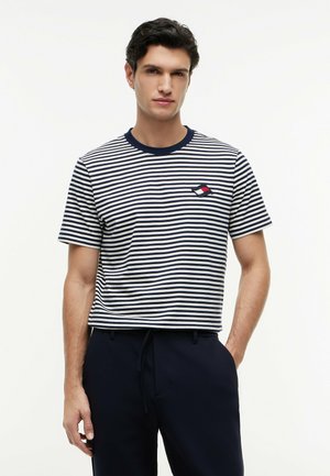 Mann trägt kurzärmeliges Shirt mit marineblauen und weißen Streifen und kleinem rot-weiß-marinblauem Logo auf der Brust sowie dunkle Hosen vor weißem Hintergrund.