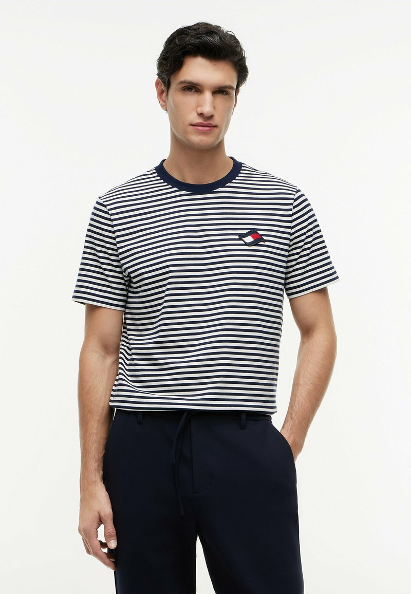 Homme portant une chemise à manches courtes rayée bleu marine et blanche avec un petit logo rouge, blanc et bleu marine sur la poitrine, et un pantalon foncé sur fond blanc.