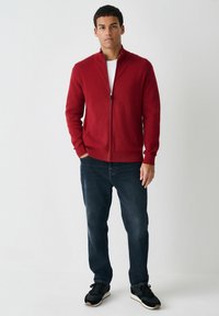 Red zip-up sweater with a ribbed collar and cuffs, paired with dark jeans and black athletic shoes. Simple, fitted design.
