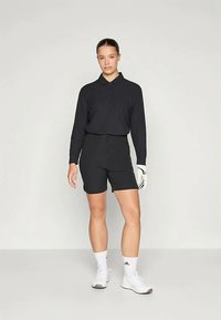 Ženska v črnem dolgih rokavih polo majici, črnih kratkih hlačah, belih športnih rokavicah na levi roki, belih nogavicah in belih supergah, ki stoji ob enobarvni ozadju.