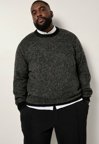 Dunkelgrauer gestrickter Pullover mit einem runden schwarzen Halsausschnitt, getragen über einem weißen Hemd, kombiniert mit schwarzen Hosen. Texturierte Stoffdetails sichtbar.
