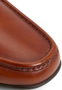 Primer plano de un zapato de cuero marrón, con textura suave, punta redondeada y costuras contrastantes a lo largo del borde. Suela de goma negra visible.