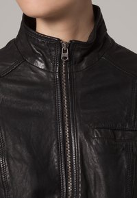 Czarna skórzana kurtka z wysokim kołnierzem, zamkiem błyskawicznym z przodu, teksturowaną powierzchnią oraz małą zewnętrzną kieszenią po lewej stronie.