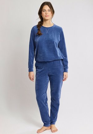 Femme debout pieds nus portant un haut bleu en velours côtelé à manches longues et un pantalon assorti sur un fond clair uni.