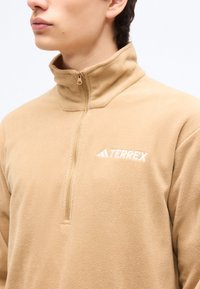 Ένας νεαρός φοράει μπεζ φλις πουλόβερ με μισό φερμουάρ και λευκό λογότυπο "TERREX" στο αριστερό στήθος, με φόντο μονόχρωμο.