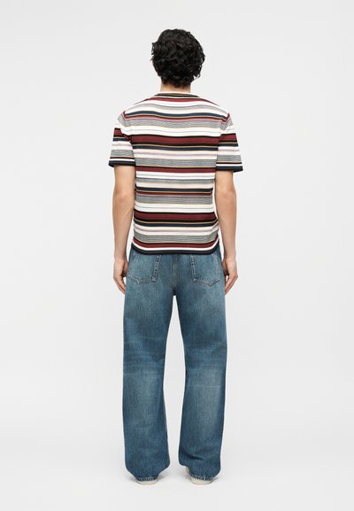 Man with curly hair wearing a short-sleeve striped shirt and loose-fitting blue jeans, standing facing away against a plain background.