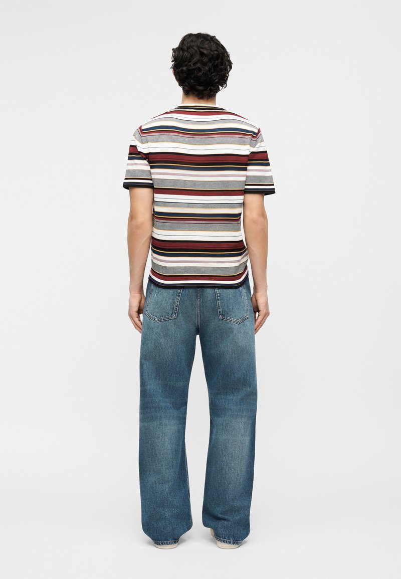 Man with curly hair wearing a short-sleeve striped shirt and loose-fitting blue jeans, standing facing away against a plain background.