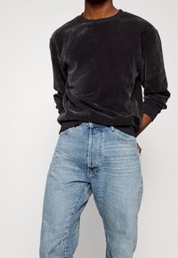 Man som bär en svart sammets långärmad topp instoppad i höga ljusblå avslappnade jeans, händerna bakom ryggen.