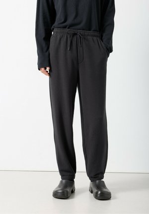 Melnas, vaļīgas jogger stila bikses ar elastīgu jostu un auklu. Faktūras audums, aproces potīšu daļā, kopā ar melnām slidena tipa kurpēm.