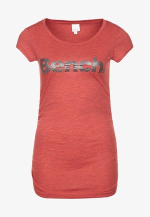 Red short-sleeve t-shirt with a rounded neckline, featuring a large silver glitter logo "Bench." on the front. Soft, textured fabric.