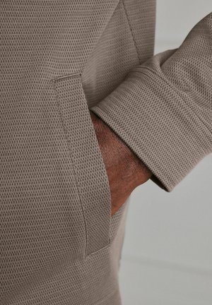 Dark-skinned hand inserted into front pocket of taupe textured long-sleeve garment against a neutral background.