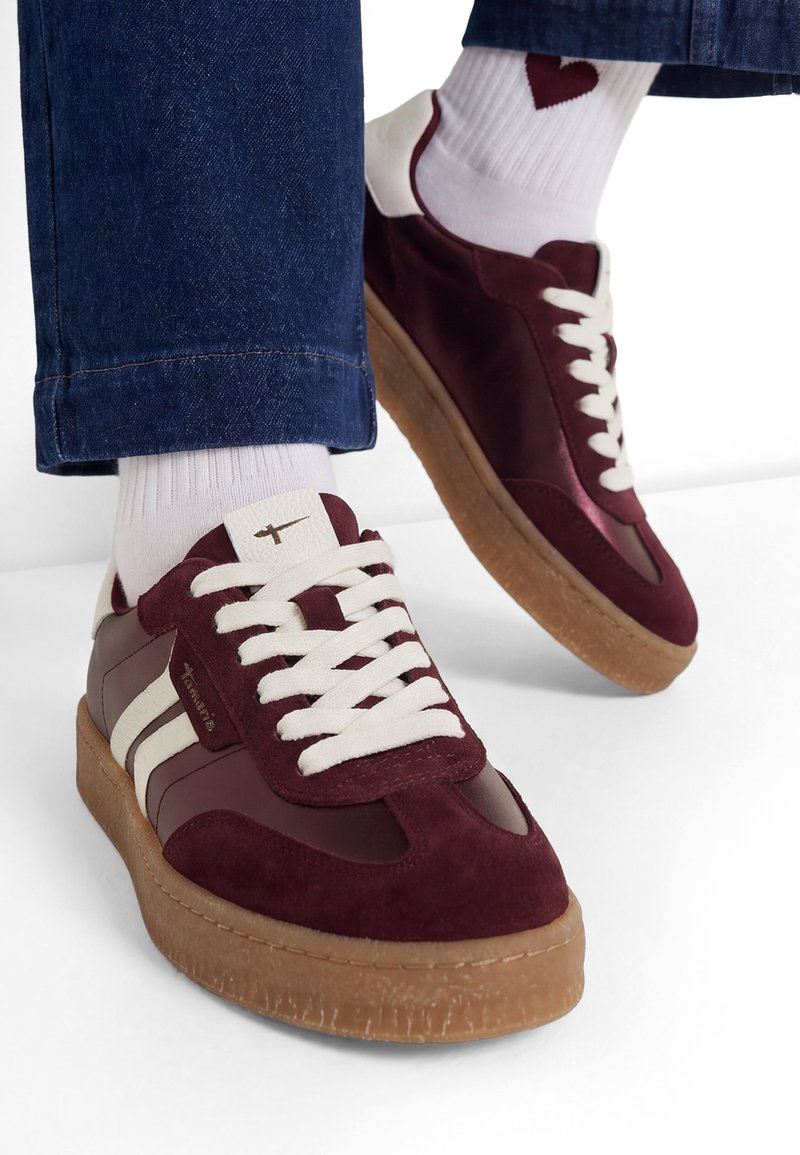 Personne portant des baskets bordeaux et blanches avec des lacets blancs, des semelles en gomme, des chaussettes blanches avec un cœur rouge et un jean bleu, debout sur un sol blanc.