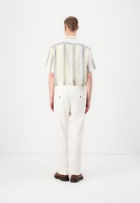 Kortärmad, randig skjorta med gula och blå linjer; kombinerad med vita, löst sittande byxor och bruna skor. Bakifrån.