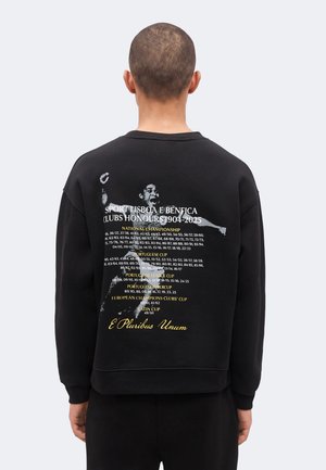 Personne portant un sweat-shirt noir avec un texte listant les honneurs du club Sport Lisboa e Benfica de 1904 à 2025 et un graphique d'un joueur célébrant.