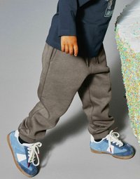 Dziecko w szarych dresowych spodniach i niebieskich sportowych butach stoi obok dużego tortu pokrytego białym lukrem i kolorowymi posypkami.