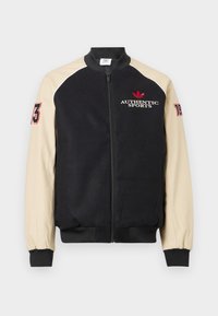 Svart og beige glidelåsjakke med "Authentic Sports" og rød logo på brystet, nummer "73" på venstre erme og "19" på høyre erme.