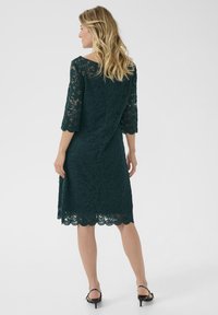 Abito in pizzo verde scuro con maniche a tre quarti, orlo scallop e silhouette aderente. La sovrapposizione di pizzo testurizzato aggiunge dettagli. Indossato con sandali con tacco.