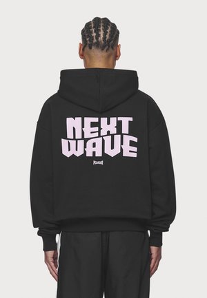 Personne avec des tresses cornrow vue de dos portant un sweat à capuche noir avec un grand texte rose "NEXT WAVE" dans le dos.