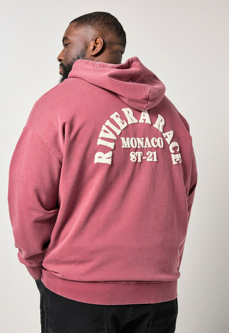 Homme tourné vers la gauche portant un sweat à capuche rouge délavé avec l'inscription "Riviera Race Monaco ST-21" imprimée au dos, sur un fond clair uni.