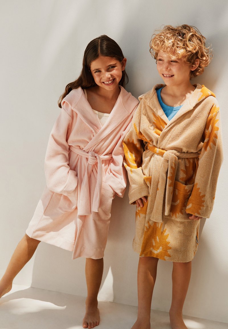 Due bambini sorridenti sono in piedi a piedi nudi contro un muro, indossando accappatoi con cappuccio; uno è rosa, l'altro indossa un accappatoio color beige con motivo a sole.