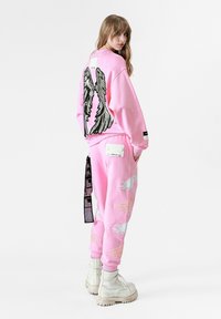 Sudadera rosa con bordado de alas de ángel negras en la espalda, combinada con joggers rosa a juego que presentan acentos de alas en blanco y dorado.