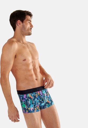 Mężczyzna bez koszulki, noszący wielokolorowe bokserki w wzory z czarnym pasem, patrzący na bok na jednolitym jasnym tle.