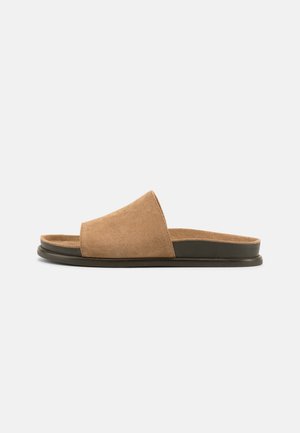 Sandale à enfiler en daim marron avec semelle noire, bout ouvert et large bride, photographiée sur un fond gris clair uni.