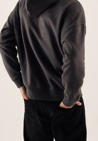 Ciemnoszara bluza z kapturem o teksturowanym wykończeniu i ściągaczach przy mankietach, zestawiona z czarnymi spodniami z tylnymi kieszeniami.